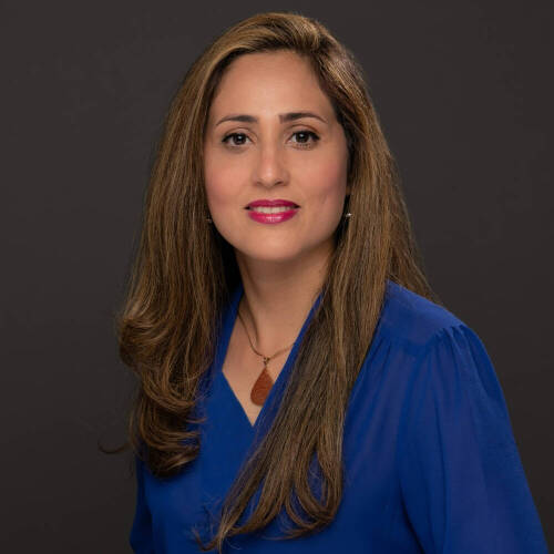 Portrait of a woman with long hair and a blue blouse, looking directly at the camera with a neutral expression. The dark gray background emphasizes her face and attire.